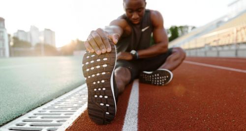 Young concentrated african male athlete stretching legs while sitting at the stadium race track